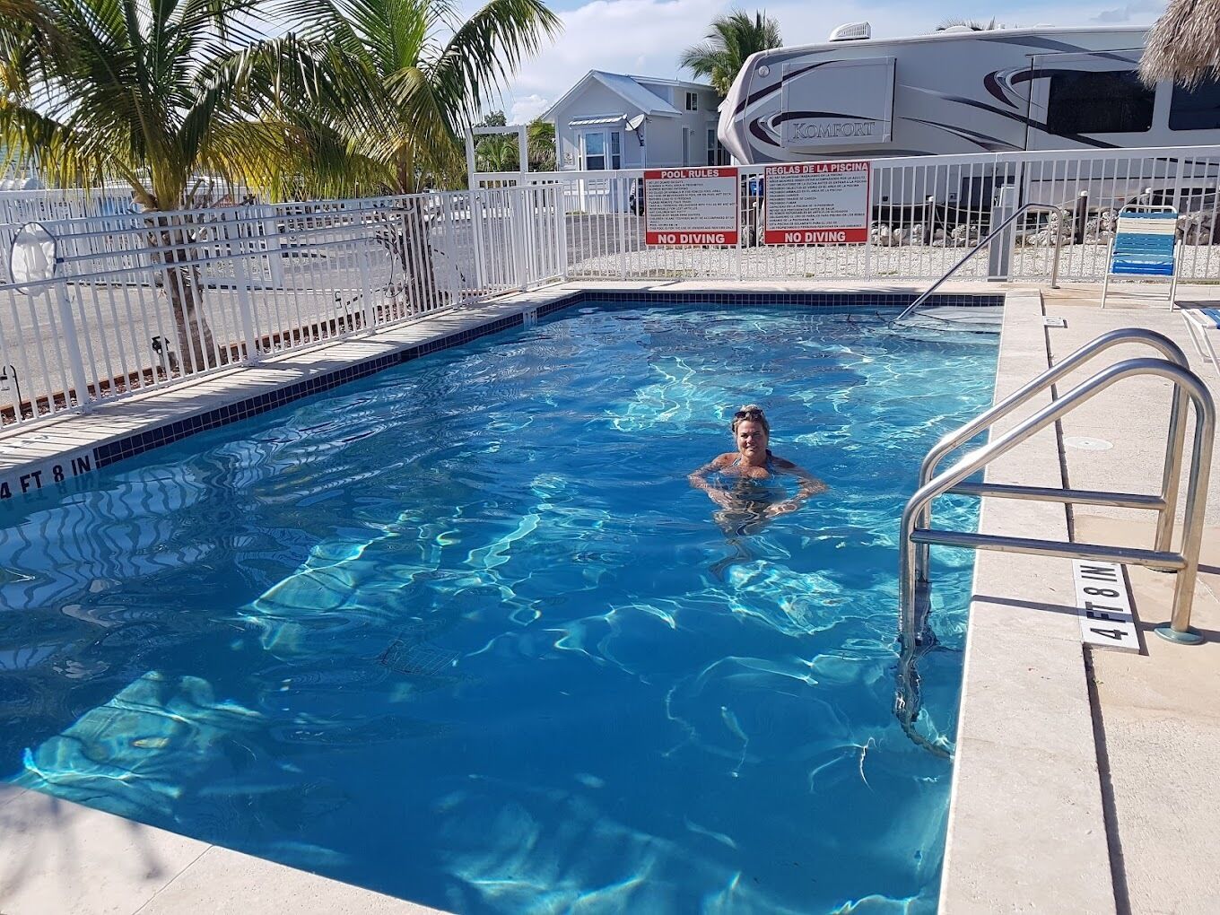 A swimming pool with a person in the water, surrounded by a white fence and palm trees.