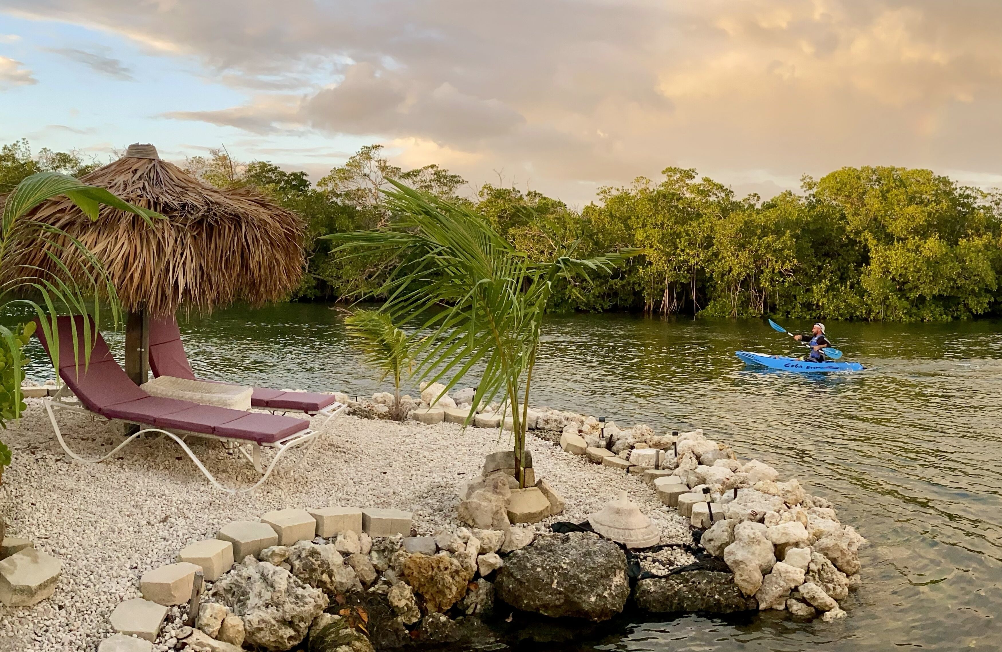 Un cadre paisible au bord de la rivière, avec un fauteuil de relaxation, une structure à toit de chaume et un petit bateau sur l’eau.