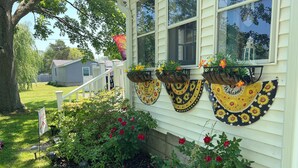 Una casa con fachada blanca, un porche con barandilla blanca y un pequeño jardín con flores rojas y un letrero.