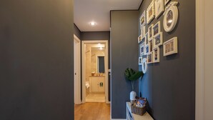 Um corredor com piso de madeira, um aparador branco com um vaso e livros, e uma parede decorada com quadros.