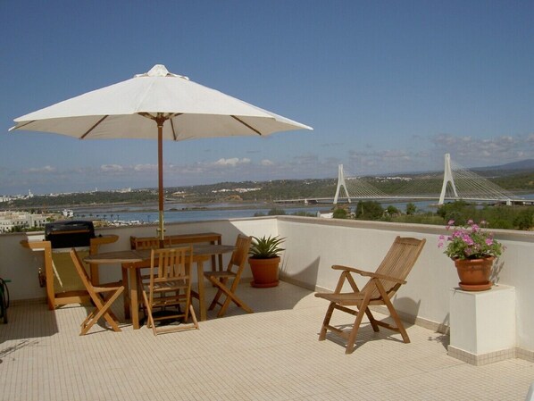 Une terrasse dotée de mobilier en bois, d’un parasol blanc et de plantes en pot.