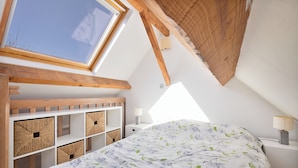Un dormitorio con tragaluces, vigas de madera, una cama con ropa de cama floral, almacenamiento empotrado y una mesita de noche con lámpara.