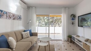 Uma sala de estar moderna com um sofá bege, uma mesa pequena branca e uma TV sobre um suporte branco.