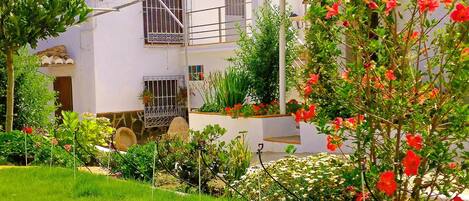 A white two-story building with a balcony, a garden with red flowers, and a clear sky.