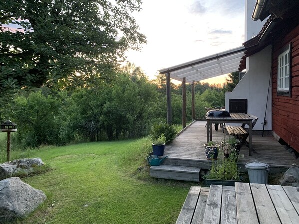 A wooden deck with a table and chairs, surrounded by greenery and a house with a red exterior.
