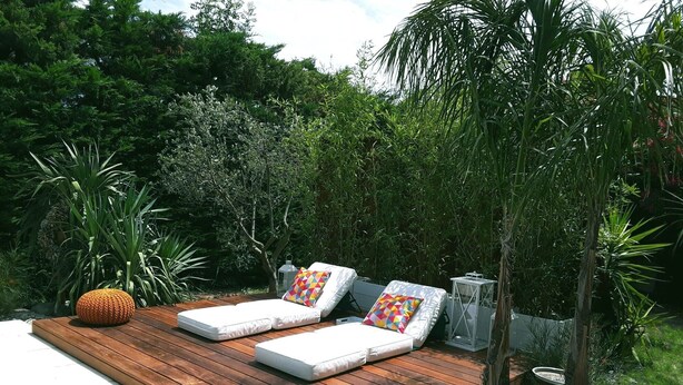 Une terrasse en bois agrémentée de deux chaises longues blanches et de coussins colorés, entourée d’une végétation luxuriante et d’une petite lanterne.