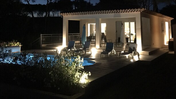 Un área de piscina moderna al aire libre con sillones de descanso y una zona de estar cubierta, rodeada de árboles y un cielo nocturno despejado.