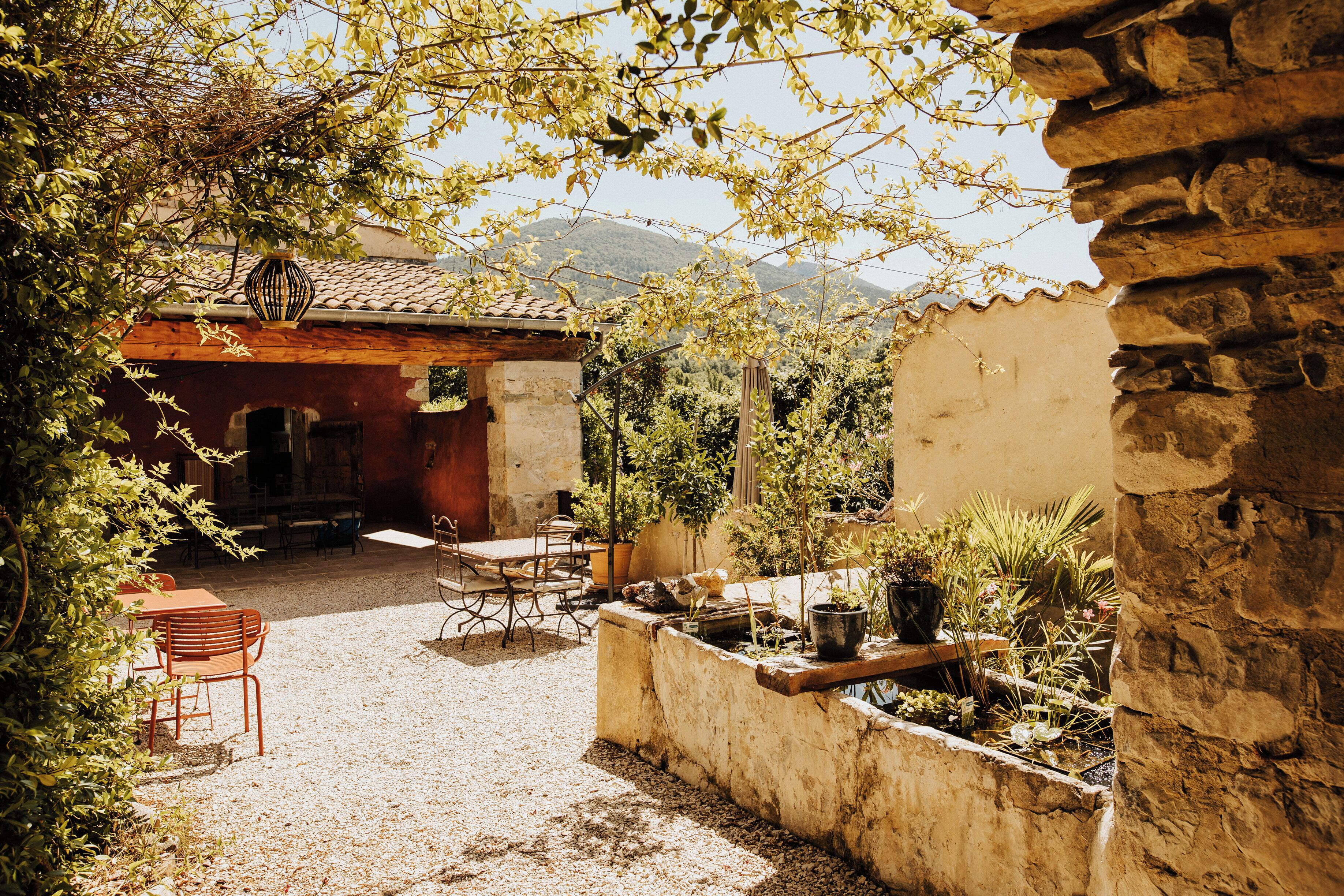 A courtyard with a rustic stone wall, a wooden table with chairs, and potted plants.