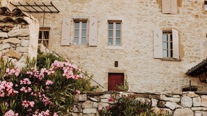 Un bâtiment en pierre avec une porte et des volets rouges, entouré d’arbustes en fleurs.