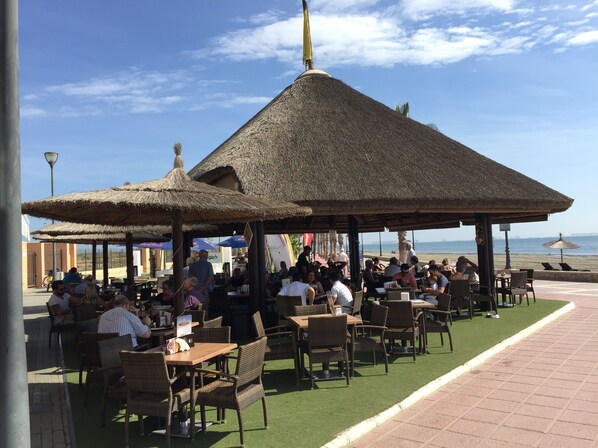 An outdoor seating area with thatched umbrellas and wicker furniture, overlooking a beach.
