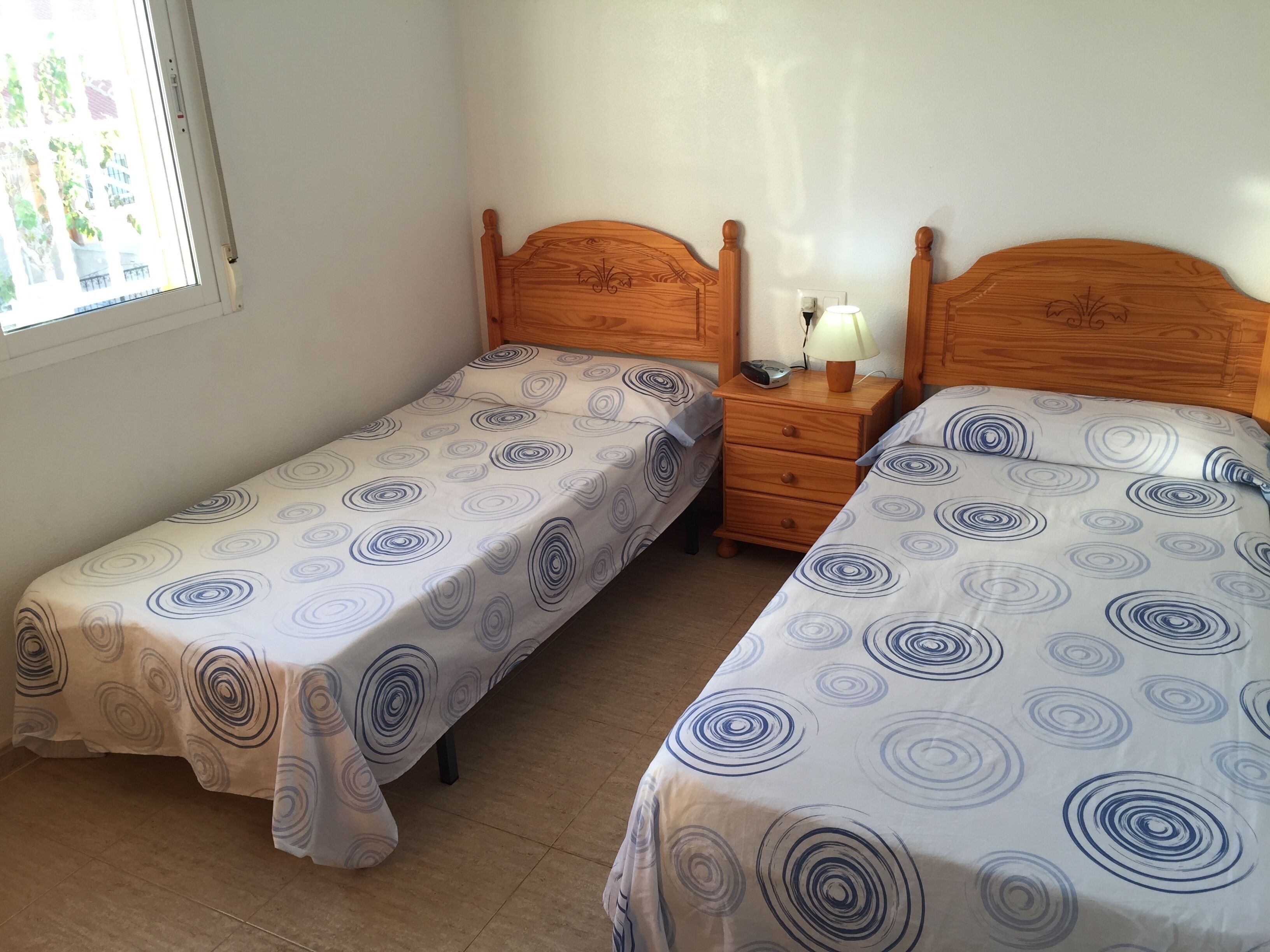 Een slaapkamer met twee eenpersoonsbedden, een nachtkastje met een lamp en een raam met uitzicht op gebouwen.