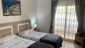 A room with two beds, a nightstand with a lamp, a painting on the wall, and a window with curtains.