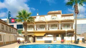 A swimming pool with a tiled deck, palm trees, and a building with balconies in the background.