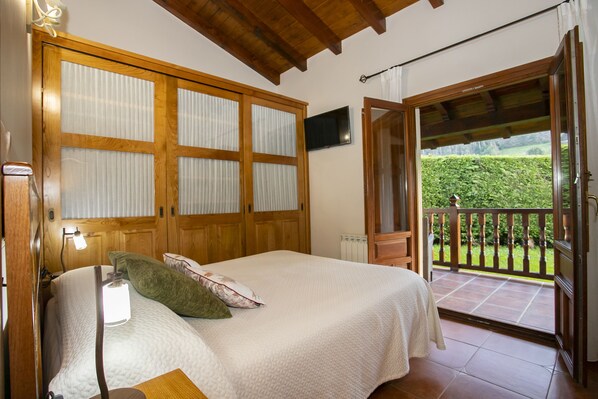 A bedroom with a bed, a nightstand, a TV, and a balcony with a view of greenery.