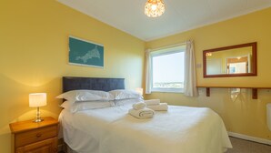 A neatly made bed with white linens, a wooden nightstand with a lamp, a framed picture on the wall, and a window with curtains.