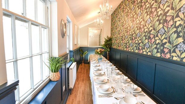 Ein langer Esstisch mit weißer Tischdecke, Tellern, Gläsern und Besteck, neben Fenstern und einer Wand mit Blumenmuster.