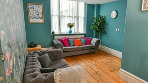 Ein Zimmer mit türkisfarbenen Wänden, einem grauen Sofa mit bunten Kissen, einem großen Fenster mit durchsichtigen Vorhängen und einer Kristallleuchter.