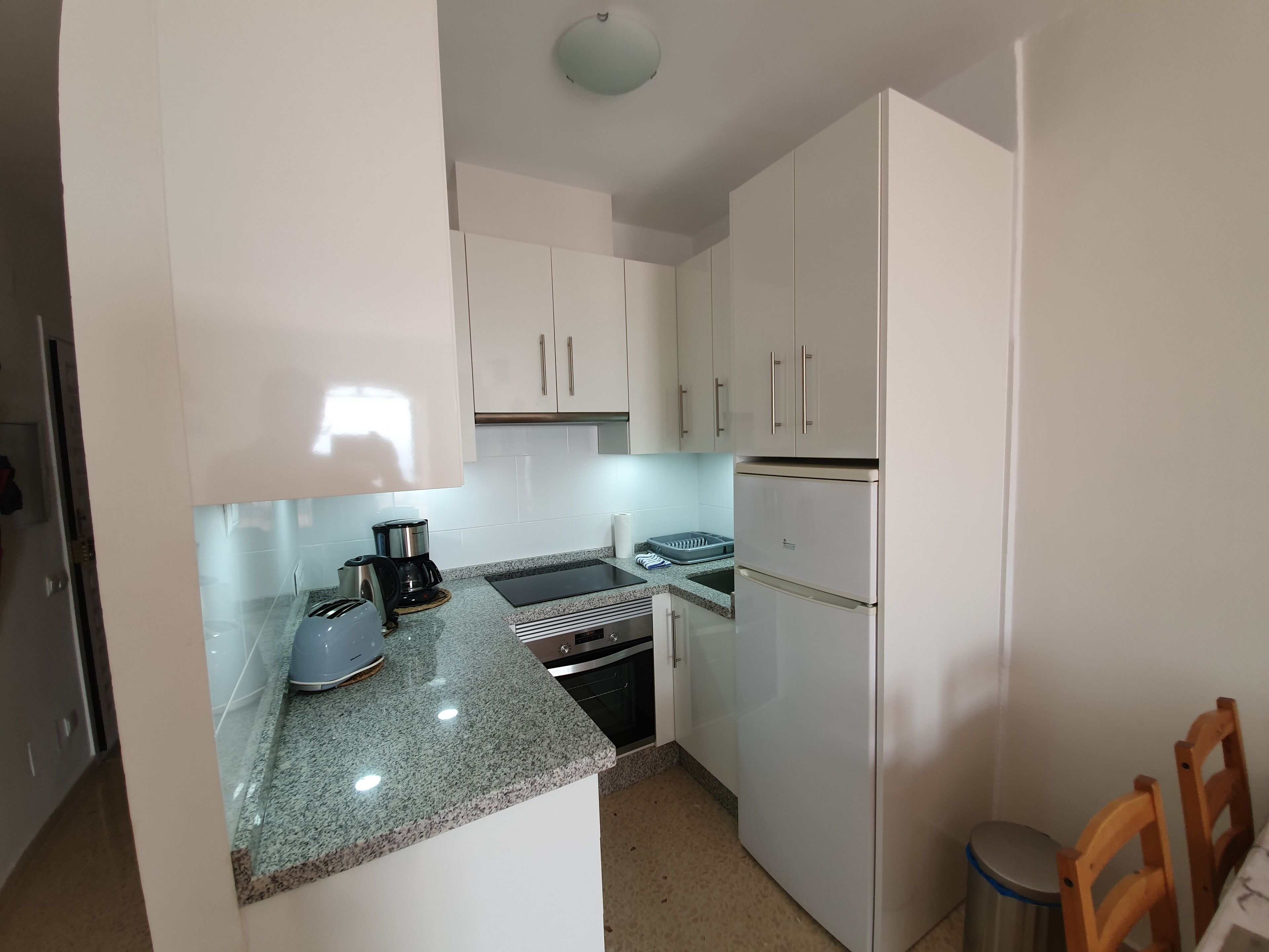 A compact kitchen with white cabinets, a built-in oven, a stainless steel refrigerator, and a granite countertop.