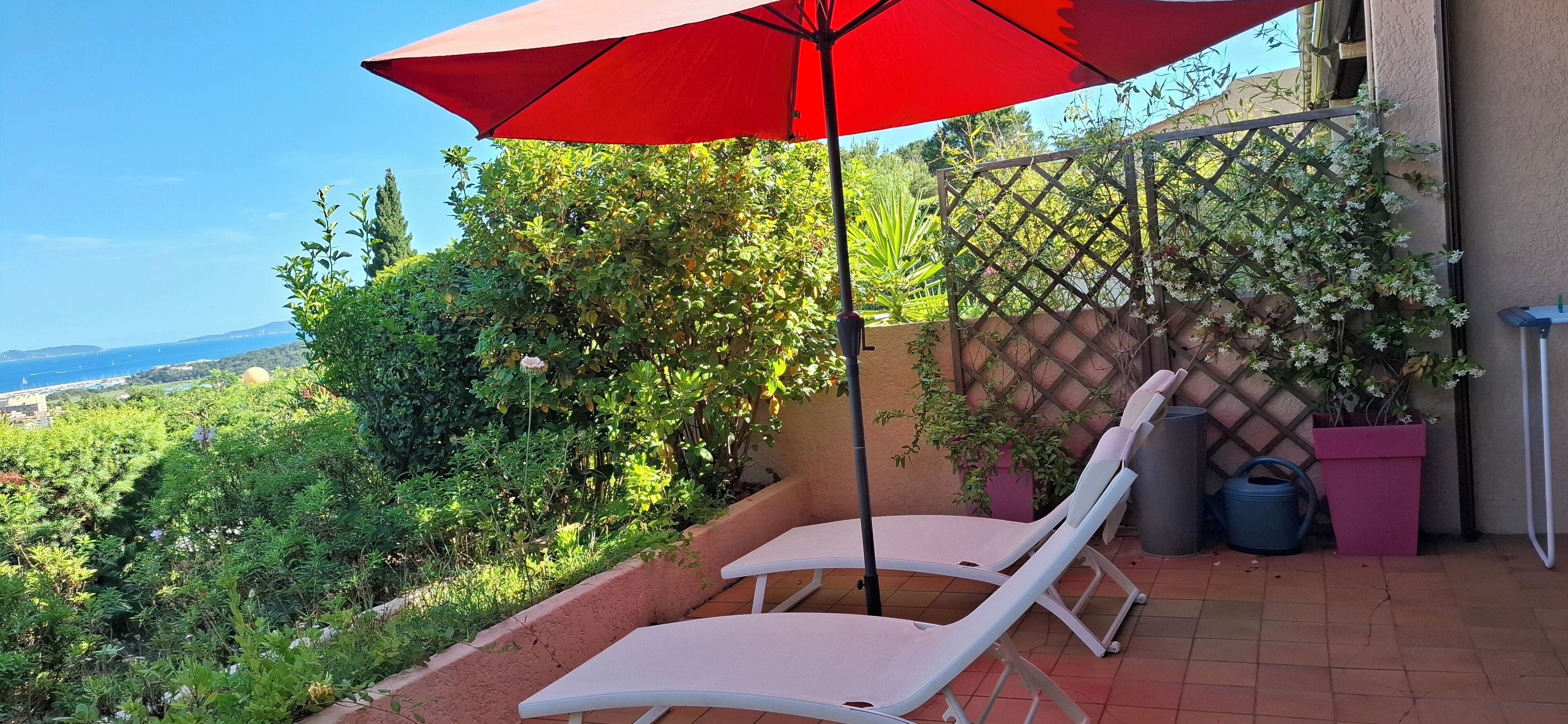 Eine Terrasse mit zwei weißen Lounge-Sesseln unter einem roten Sonnenschirm, umgeben von üppigem Grün und Blick aufs Meer.