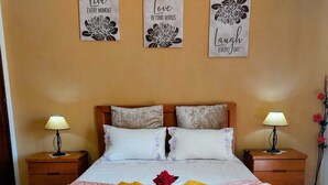 Una habitación de hotel con una cama, dos mesitas de noche, una lámpara de noche, una flor decorativa y tres obras de arte en la pared.