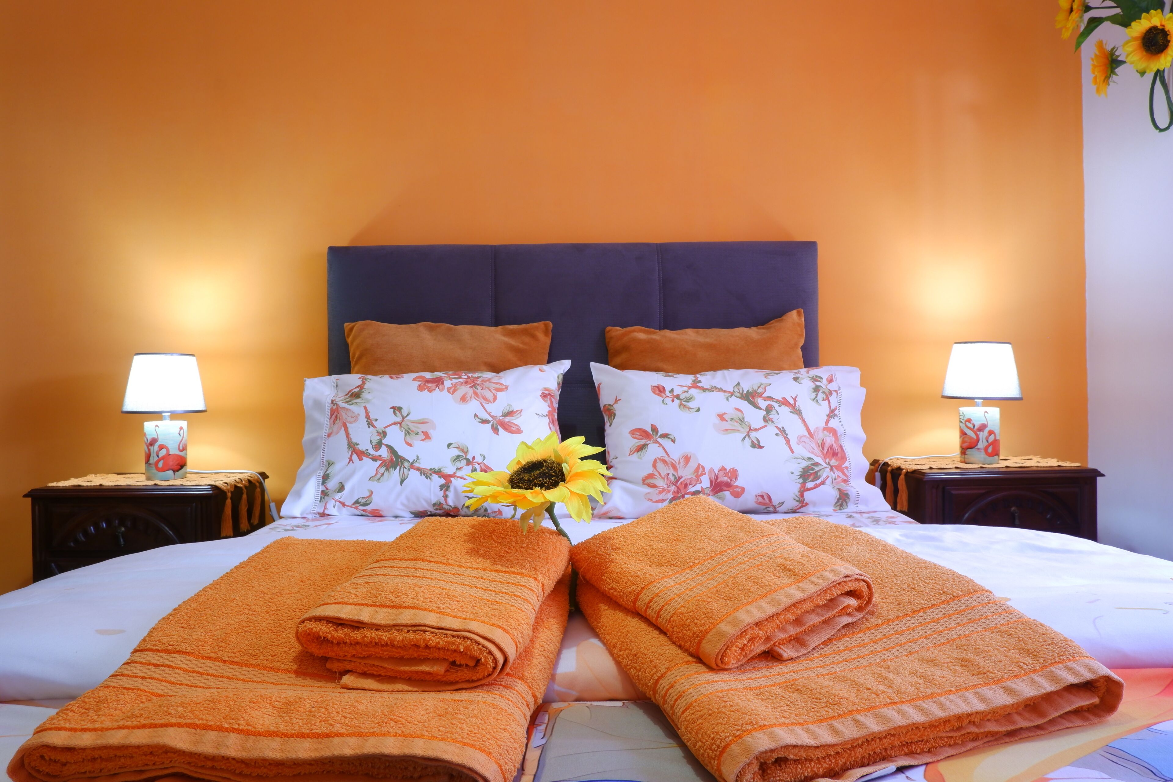A neatly made bed with orange towels, floral pillows, and a sunflower decoration.