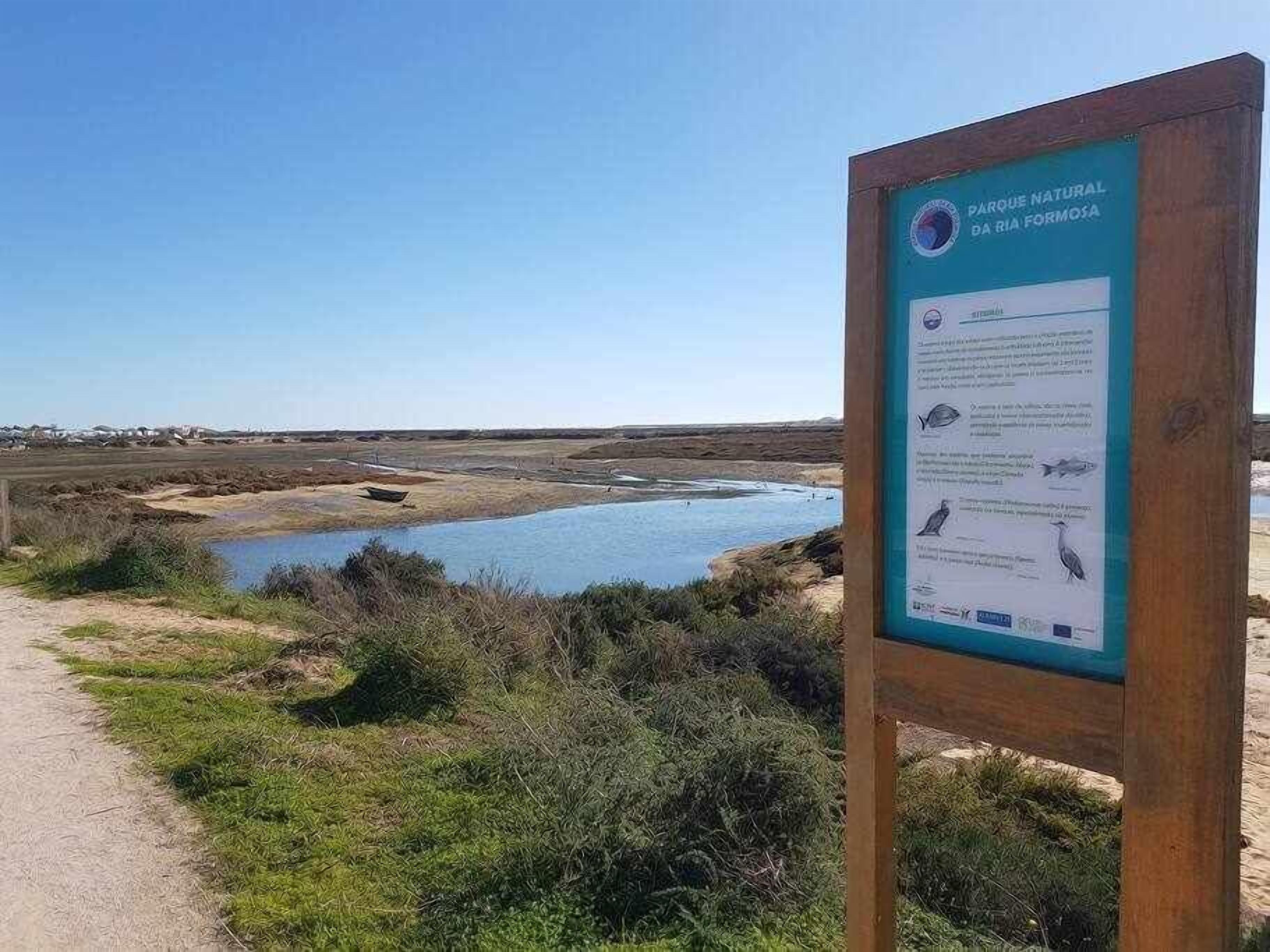 Cartell del Parc Natural de la Ria Formosa amb un paisatge costaner al fons.