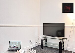 Une chambre moderne avec un téléviseur à écran plat posé sur un support en verre, un ordinateur portable sur une petite table et une enseigne « Klasse Living » accrochée au mur.