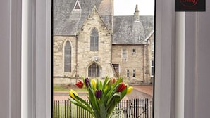 Une vue d’un bâtiment en pierre, doté d’un toit en pente et de fenêtres en arc de cercle, prise à travers une porte surmontée d’une grille en métal noir au premier plan.
