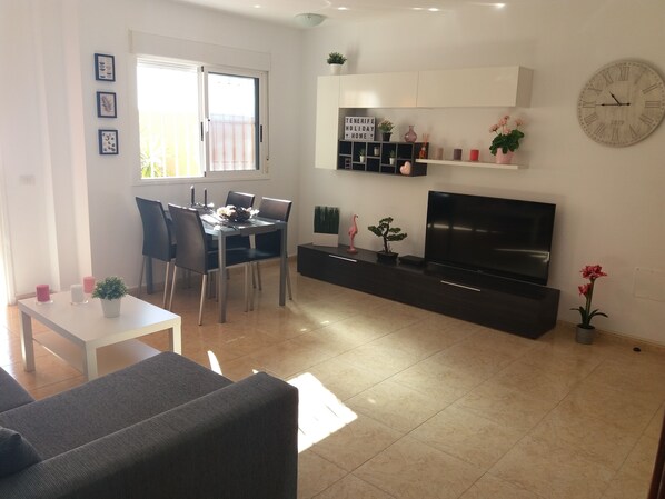 A modern living room with a dining area, a flat-screen TV, and a large clock on the wall.