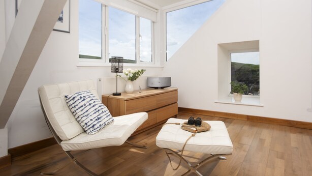 A modern loft bedroom with a sloped ceiling, large windows, a bed, a seating area, and a wooden dresser. - A beautiful, double-fronted terrace house situated in Port Isaac. (Port Isaac)