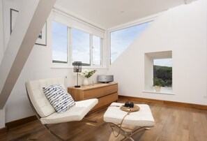 A modern loft bedroom with a sloped ceiling, large windows, a bed, a seating area, and a wooden dresser.