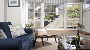 A room with a blue sofa, a wicker chair, a dining table with wine glasses and a bottle, and a bay window with a view of a stone house and greenery. - A beautiful, double-fronted terrace house situated in Port Isaac. (Port Isaac)