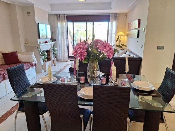 Un comedor con una mesa de vidrio dispuesta para cuatro personas, rodeada de sillas oscuras. Un jarrón con flores rosadas está sobre la mesa, y hay velas y vasos.