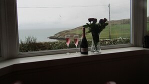 Ett fönster med utsikt över havet och kullarna, två glas vin och en vas med blommor på fönsterbrädan.
