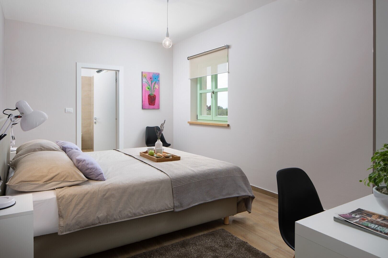 Een slaapkamer met een bed, nachtkastje, lamp, stoel, bureau, raam en kunstwerk.