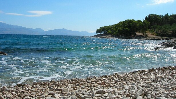 Ein felsiger Strand mit klarem, blauem Wasser und Bergen im Hintergrund.