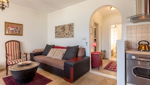 Ein Wohnzimmer mit einer Couch, einem Sessel und einem Couchtisch, eine Küche mit Backofen und Wasserkocher sowie ein Flur, der zu einem weiteren Zimmer führt.