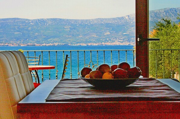 A balcony with a table, a bowl of fruit, and a view of the sea and mountains.
