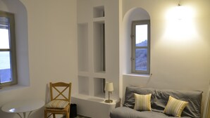 A room with a grey sofa, a wooden chair, a small round table, and a lamp. There are two windows with views of the outside.