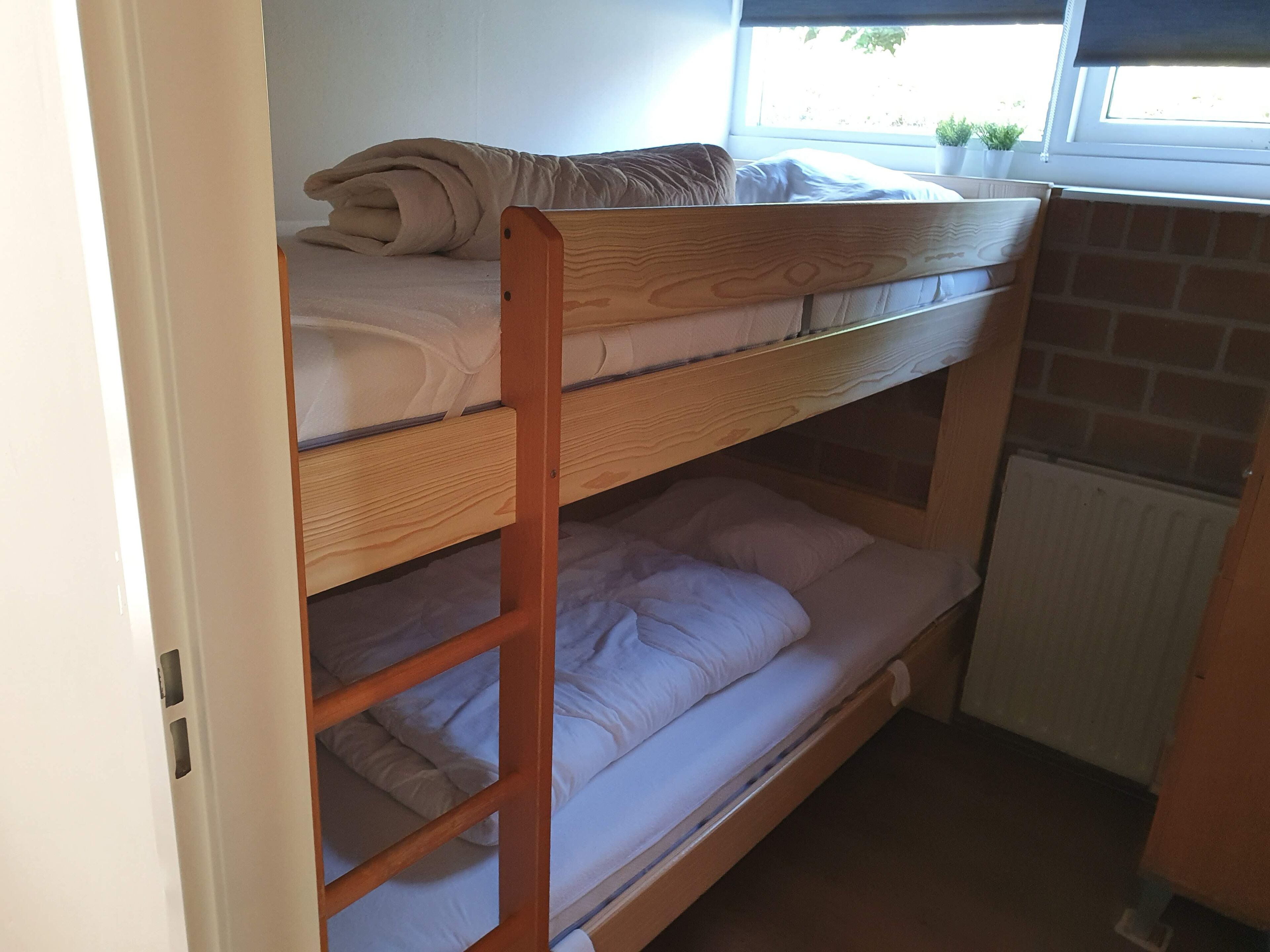 A bunk bed with a wooden frame, a window with a view of greenery, and a radiator below the lower bunk.