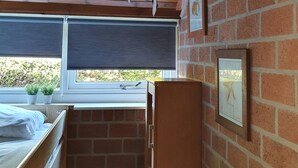 Una habitación con techo de madera, pared de ladrillo, ventana con vistas a la vegetación, cama, radiador y un cuadro enmarcado.