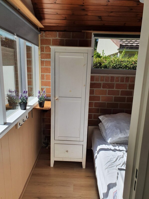 Una habitación pequeña con techo de madera, un armario blanco, una cama con ropa de cama blanca, una ventana que da a zonas verdes y un estante con plantas en macetas.