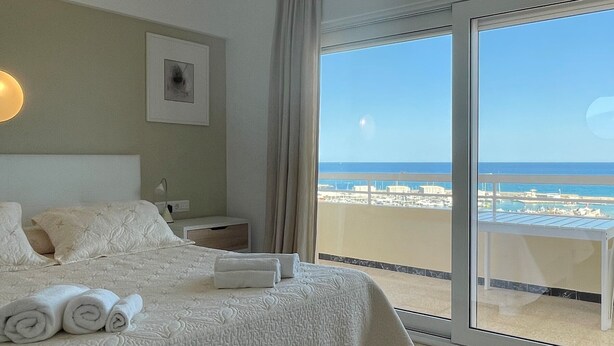 A hotel room with a bed, a nightstand, a lamp, a framed picture, and a balcony with a view of the beach and ocean. - Beautiful sea view from all rooms, communal swimming pool / tennis (El Campello)