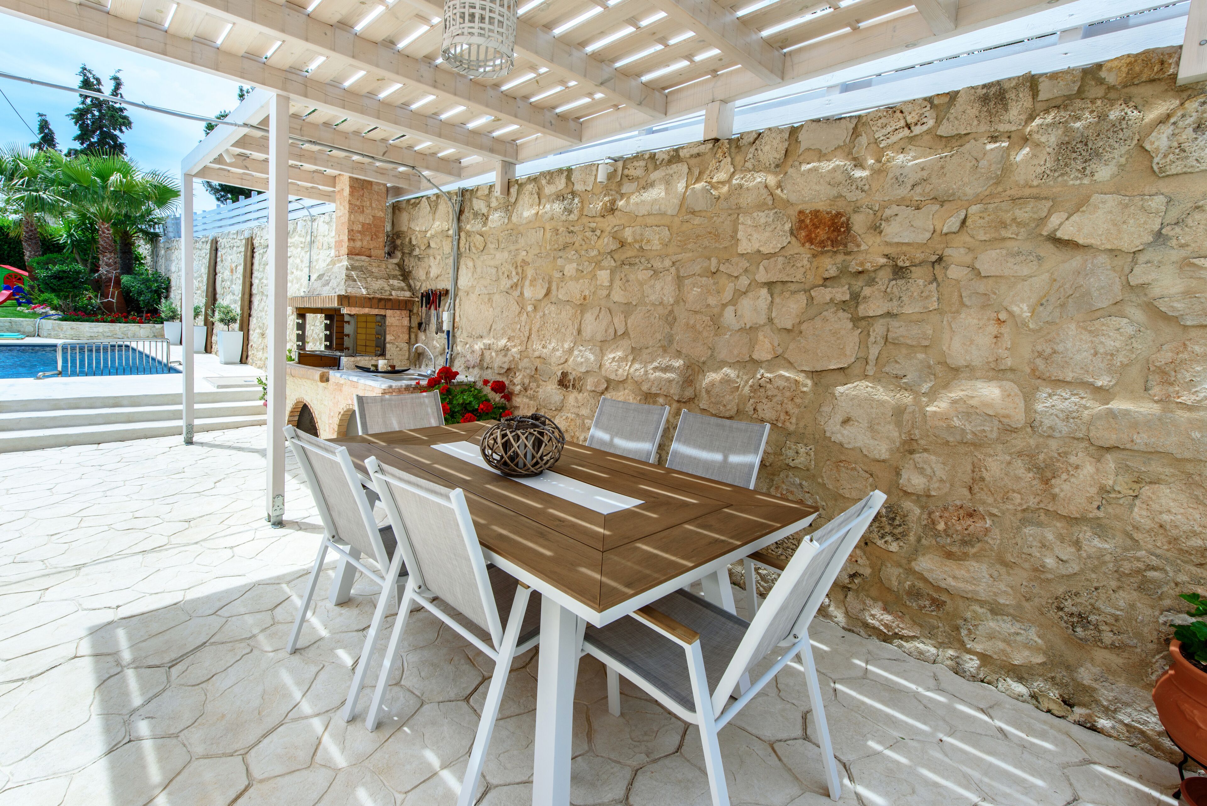 An outdoor patio with a stone wall, a wooden table with chairs, and a pool in the background.