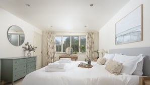 A bedroom with a large bed, a dresser, a mirror, a window with curtains, and a painting on the wall. - The Nineteenth is a traditional four bedroom house overlooking Roserrow lakes. (Wadebridge)