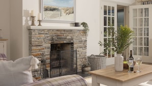 A cozy living room with a stone fireplace, a painting above it, and a dining area with a table set for two. - The Nineteenth is a traditional four bedroom house overlooking Roserrow lakes. (Wadebridge)