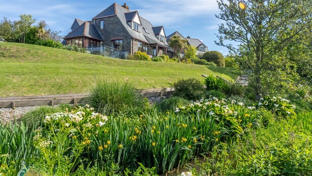 A large house with multiple roofs, surrounded by lush greenery and a garden with various plants and flowers. - The Nineteenth is a traditional four bedroom house overlooking Roserrow lakes. (Wadebridge)