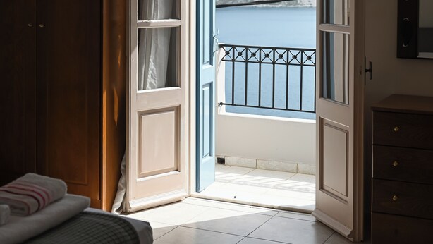 Ein Zimmer mit einem Bett, einem Kleiderschrank, einer offenen Tür, die auf einen Balkon mit Meer- und Bergblick führt.