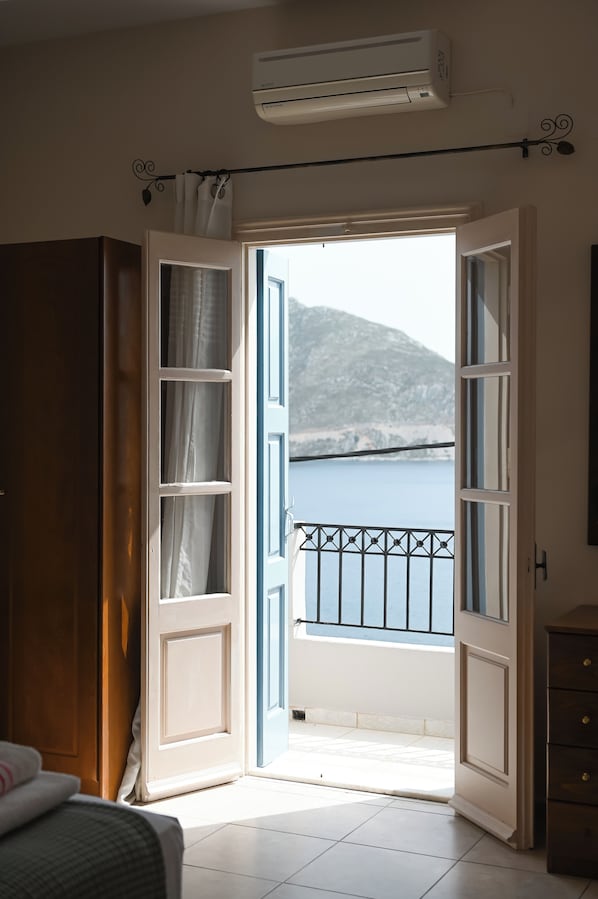 Ein Zimmer mit offener Tür, das auf einen Balkon mit Blick auf ein Gewässer und ein Gebirge führt.