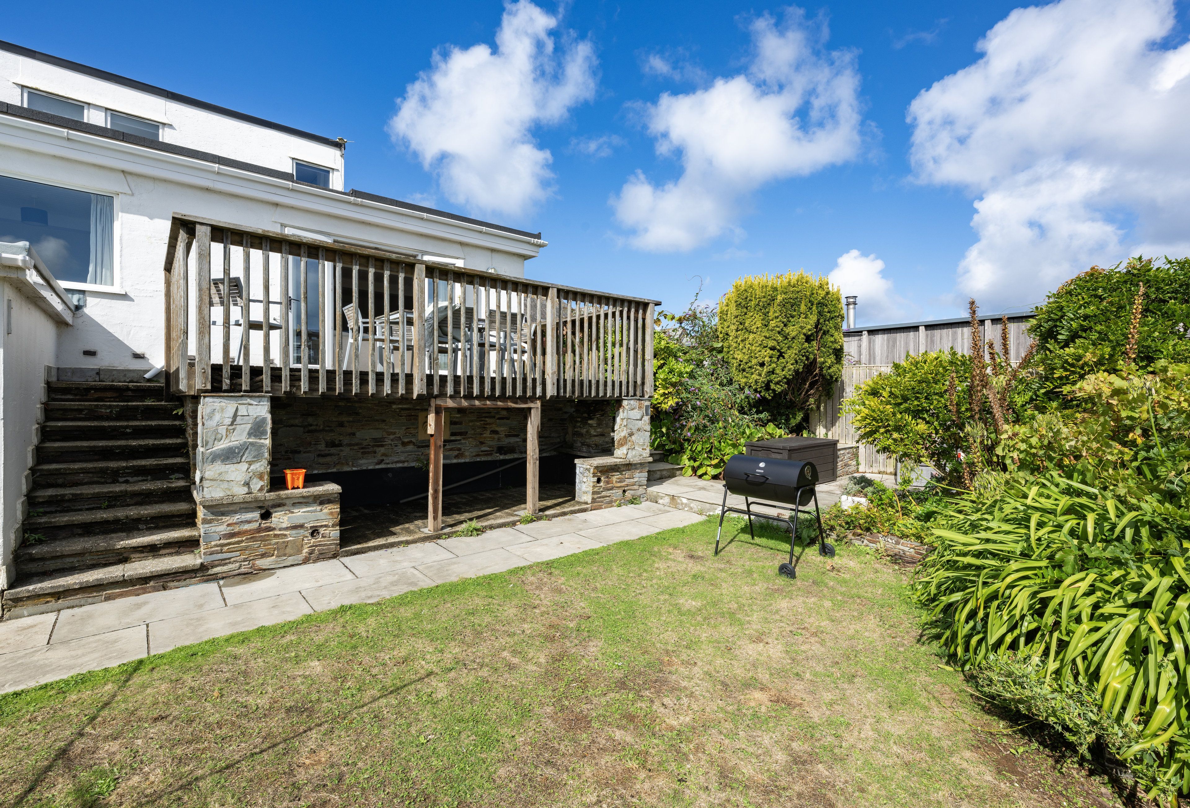 A modern house with a wooden deck, a stone staircase, and a barbecue grill in the yard.